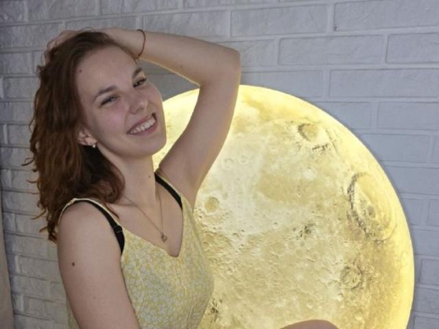 Chaya Parrett mit lockigem, braunem Haar, lächelnd, mit einer Hand im Haar, trägt ein gelbes, gemustertes Kleid und steht vor einem leuchtenden Vollmond. Sie hat kleine Brüste und ein süßes Gesicht..