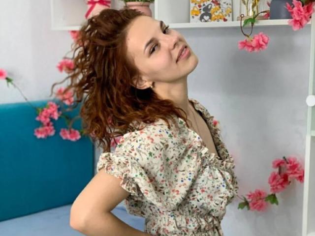 ChayaParrett with wavy brown hair, floral dress, and tilted head, showing her small tits and cute face, surrounded by pink flowers and white shelves.