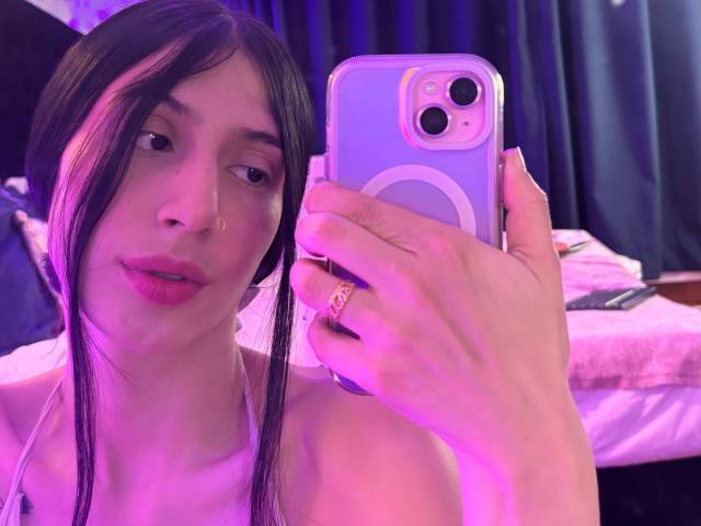 Sophie Beaumont, met lang zwart haar, een neusring en roze lippen, houdt een paarse telefoon vast en maakt een selfie. Ze draagt een ring en de belichting is roze en paars. De borsten zijn niet zichtbaar..