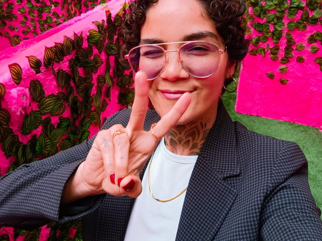 AnaRoa met krullend haar, een roze bril en rode nagellak, die een vinger op haar lippen houdt, draagt een geruit blazer, een wit shirt en een gouden ketting..