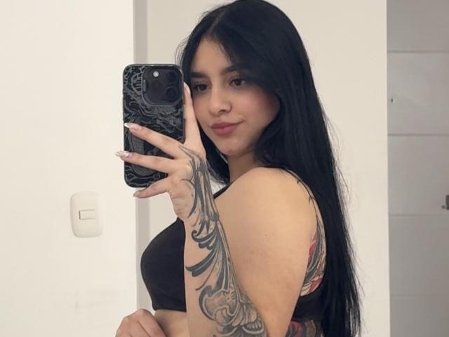 MeganPaige, con cabello largo y negro, pequeños senos en un top deportivo negro, sosteniendo un teléfono con una funda negra, un tatuaje en su brazo derecho, sonriendo a la cámara..