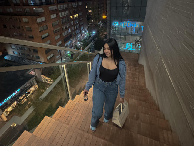 MeganPaige with big ass and tits, wearing a black top, blue jeans, and a jacket, holding a bag and phone, walking down a brick staircase at night.