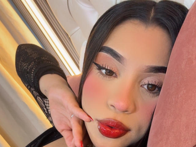 LunaVali has red lipstick, glossy pink eyes, and dark eyebrows. She's touching her cheek with a red-manicured finger, wearing a black lace long-sleeve top.
