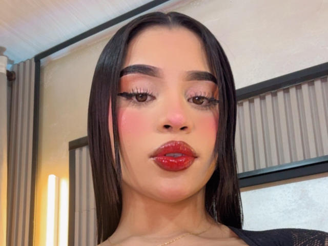 LunaVali has shiny black hair, pink blushed cheeks, red glossy lips, and long eyelashes, looking straight at the camera with a serious expression. Her eyebrows are thick and dark.