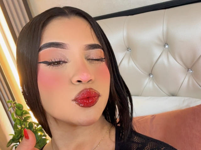 LunaVali, com lábios vermelhos brilhantes, blush rosa e delineador preto marcante, fazendo um leve biquinho com os olhos fechados, cabelos pretos, e vestindo uma blusa de malha preta, está em pé ao lado de uma cabeceira estofada..
