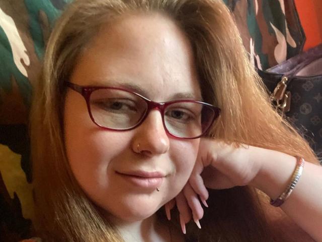 NymphoPrincessBi - ragazza attraente con occhiali rossi, capelli castano chiaro, piercing al naso, anello al labbro, sorriso appena accennato, mano appoggiata sulla testa, braccialetto al polso, viso leggermente paffuto, espressione rilassata..