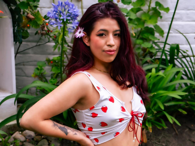 ElizaNior with dark red hair, wearing a white crop top with red heart patterns, showing her small tits, a pink flower in her hair, and a tattoo on her arm.