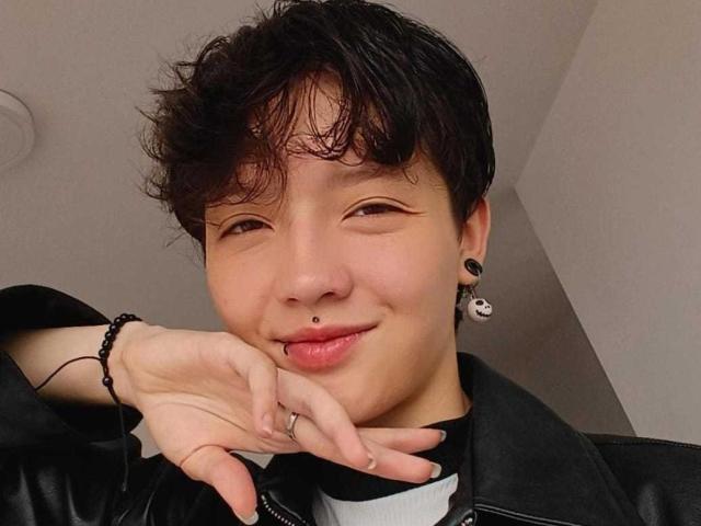 LissyLuxe has short, messy black hair, a nose piercing, and black ear studs. She's smiling with a hand on her chin, wearing a black leather jacket.