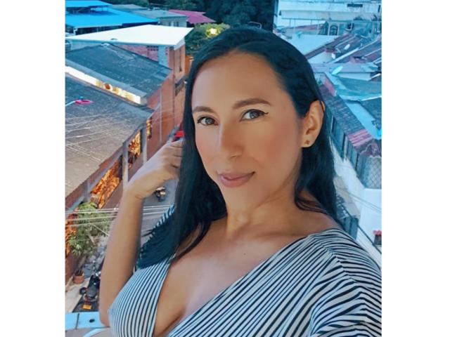 Jade Foster, avec ses cheveux noirs, son haut rayé et sa poitrine décolletée, sourit à la caméra, une main dans ses cheveux, des boucles d'oreilles visibles, un ciel bleu en arrière-plan, un léger décor de toit urbain..