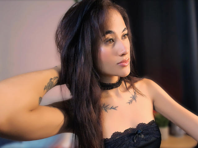HarleyRoods con cabello negro largo, collarín negro, tatuajes en el pecho y un top de encaje negro sin tirantes, mirando hacia un lado, con la mano en el cabello, ojos marrones, piel clara y labios carnosos..