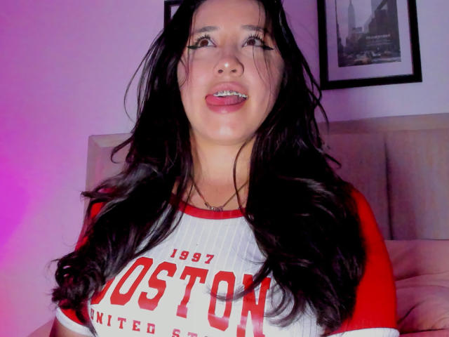 JessyBarnnett a de longs cheveux noirs, une chemise raglan rouge et blanche, et elle tire la langue en montrant ses dents en métal, elle a l'air mignonne et séduisante..