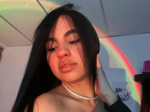 Dalila Tennesse, con i suoi lunghi capelli neri, gli occhi chiusi e una collana di perle, ha un riflesso arcobaleno sul viso, una mano che tocca i capelli e un sorriso appena accennato sulle sue labbra lucide..