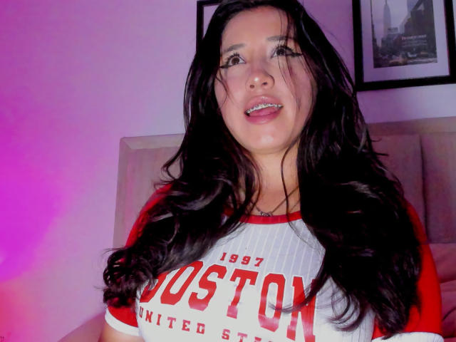 JessyBarnnett has long black hair, red and white baseball shirt, and is smiling with her tongue slightly out, showing her teeth. Pink and purple lights are in the background.