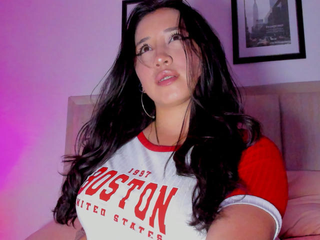JessyBarnnett, avec de longs cheveux noirs, un t-shirt rouge et blanc "Boston United States", des boucles d'oreilles cercles, des lèvres légèrement entrouvertes de couleur rose, un éclairage rose-violet, regardant sur le côté, un lit en arrière-plan..