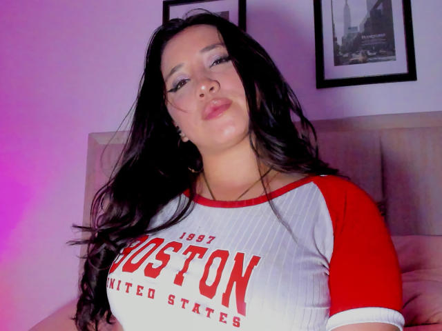 JessyBarnnett, avec de longs cheveux noirs, des lèvres pulpeuses, portant un t-shirt rouge et blanc "1997 Boston United States", montrant de grandes seins, assise sur un canapé beige, éclairée par une lumière violette..