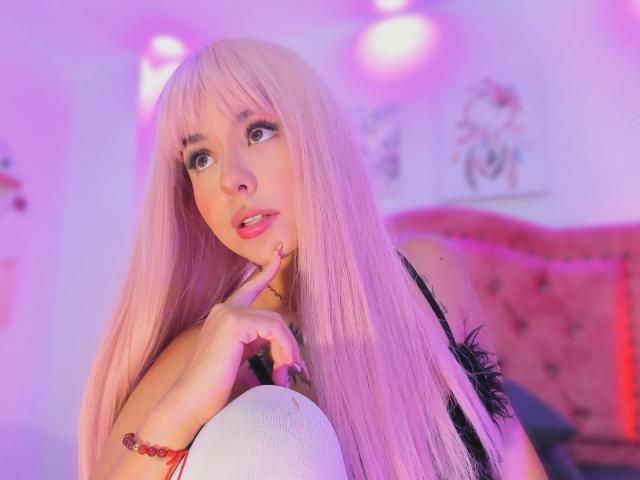 GraceClarke with pink hair, finger on lips, looking up, wearing black lingerie, pink and purple lighting, white pillow, red bracelet, and pink headboard in the background.