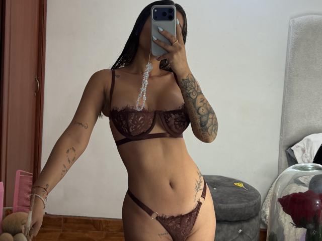 ThaliaAres en lingerie en dentelle marron, montrant ses seins et ses parties intimes dans un selfie pris dans un miroir, avec des tatouages sur les bras, tenant un téléphone, avec de longs cheveux noirs, debout dans une pièce avec une chaise..