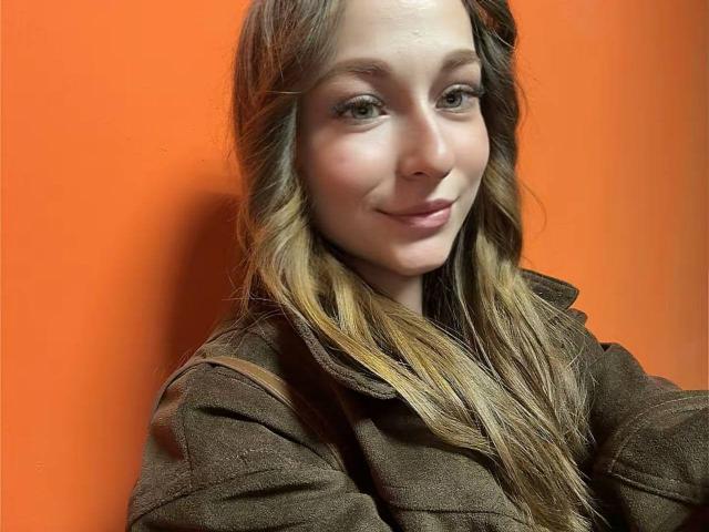 CherelleDelozier with long, wavy brown hair, wearing a brown jacket, smiling subtly, has light skin and green eyes, against an orange background.