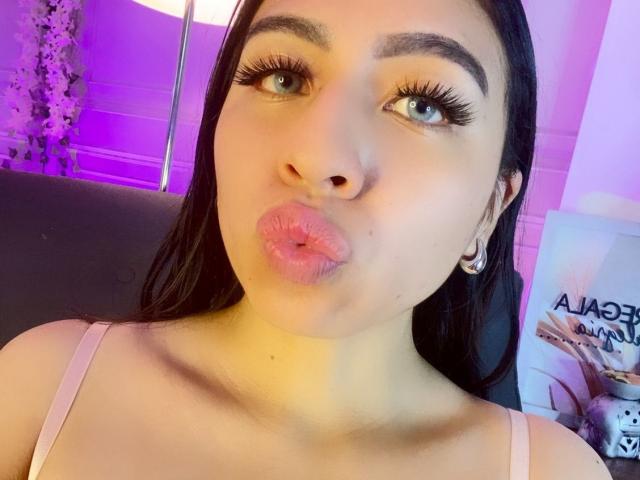 ZoeRedds has pink pouty lips, thick black eyelashes, and dark brown hair, wearing a pink top with small breasts, and a hoop earring, in a purple-lit setting.
