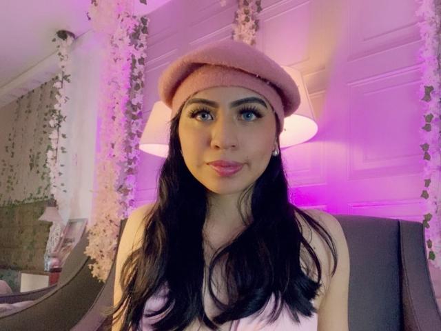 ZoeRedds, con lunghi capelli neri, un berretto rosa e rossetto rosa, sorride alla fotocamera, mostrando il seno in una canotta bianca, seduta in una stanza illuminata di viola..