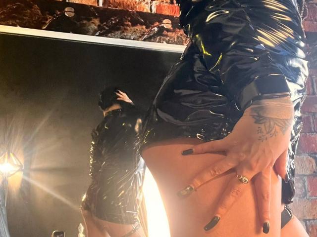 DemiMistral in shiny black latex, reflecting in a mirror. Her hand, with tattoos and black nails, rests on her bare ass. She's wearing a black mask.