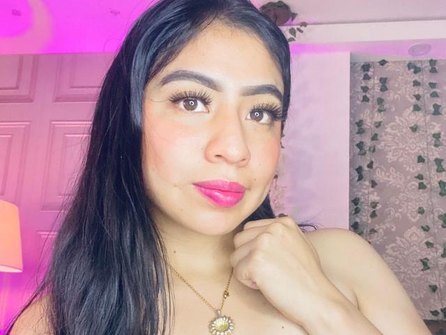 ZoeRedds has long black hair, pink lipstick, and a gold necklace with a sun pendant, holding her chin with her hand, showing off her big brown eyes and cute face.