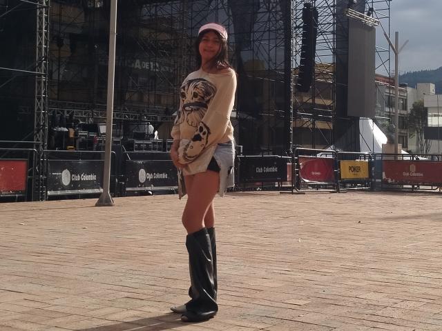 VivienSandrina stands with a smile, wearing a pink cap, beige long-sleeve shirt with a tiger print, denim shorts, and black knee-high boots, in a sunlit urban area.