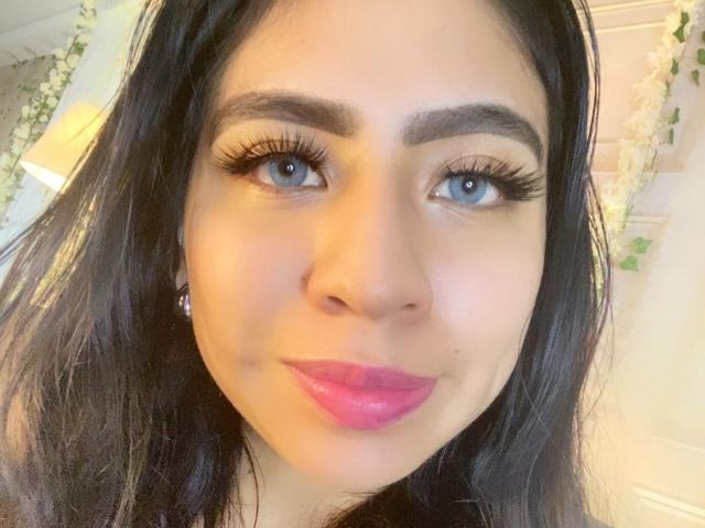 ZoeRedds has blue eyes, thick black eyebrows, long black eyelashes, pink lipstick, and dark wavy hair. She's wearing diamond earrings and smiling at the camera.