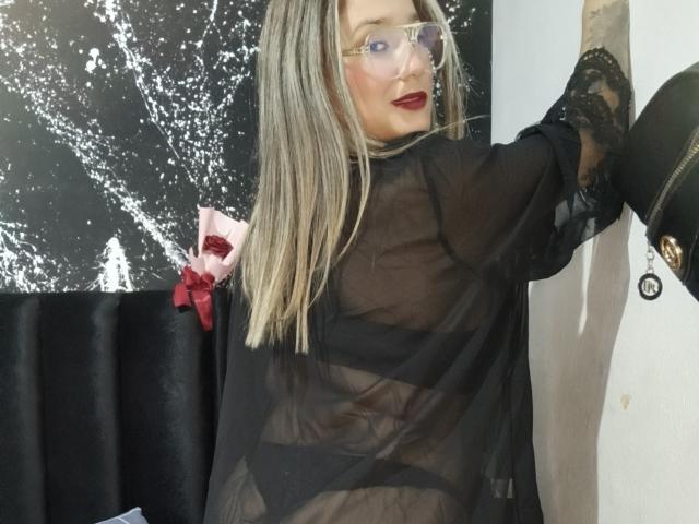 AlexaHotSeins mit blonden Haaren, rotem Lippenstift, klaren Brillen, schwarzem, durchsichtigen Oberteil, sichtbare Genitalien, schwarzen Spitzenhandschuhen, lehnt an einer Wand, schwarz-weißer Hintergrund, rotes Band auf einem schwarzen Stuhl..
