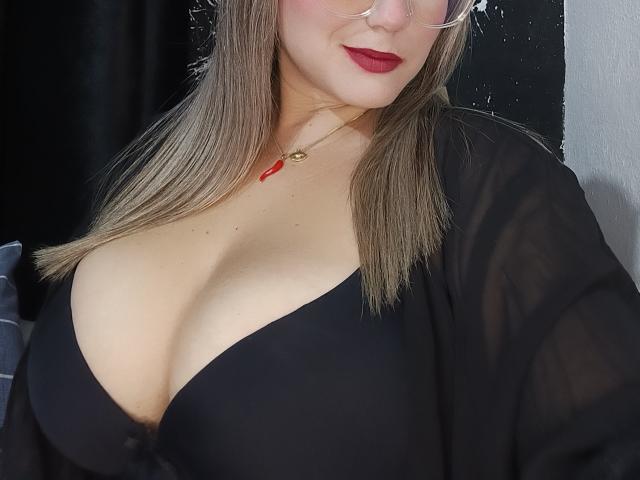 AlexaHotSeins with big tits, wearing a black bra, sheer black top, red lipstick, glasses, and a red necklace, has blonde hair and fair skin, looking down.