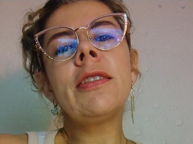 EmiliaMilf with blue eye makeup, glasses, and a slight smirk, has a wet chin and mouth, wearing a white tank top, with droplets on her face.
