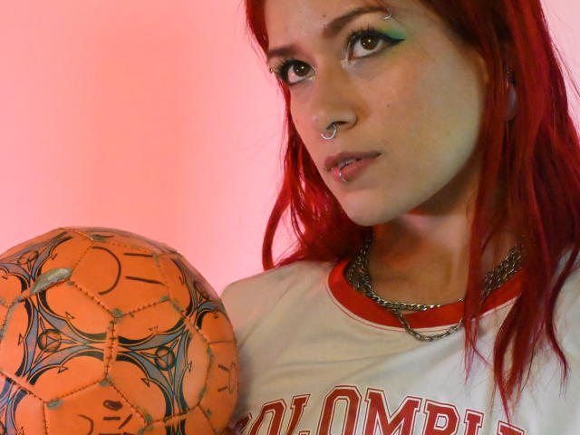 LolyYunno, con i capelli rossi, il trucco verde sugli occhi, un anello al naso e un piercing al labbro, che tiene un pallone da calcio arancione con tatuaggi neri, indossa una maglietta sportiva bianca..