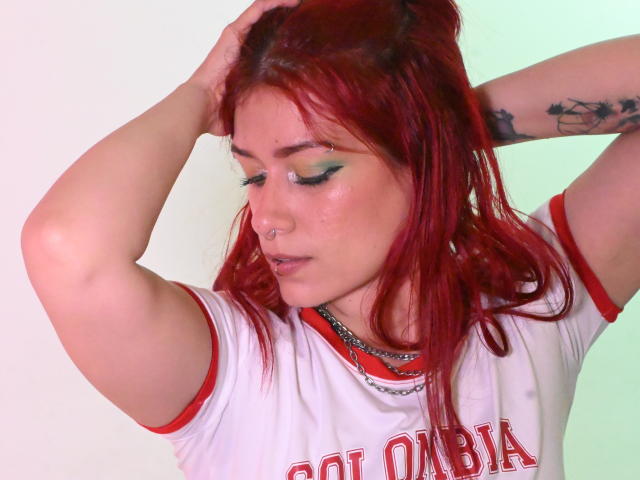 LolyYunno, avec des cheveux roux, du maquillage vert pour les yeux, un anneau au nez et un tatouage, portant un t-shirt de sport blanc avec des détails rouges, les bras levés, les mains dans les cheveux, regardant vers le bas, une lumière verte subtile..