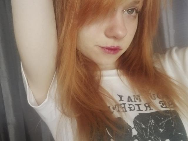 JamieLathrum, met rood haar, bleke huid en roze lippen, draagt een wit shirt, heft een arm omhoog, laat een kleine neuspiercing zien en kijkt naar de camera..