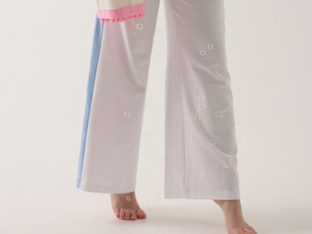 Dreia steht mit nackten Füßen in weißen Hosen mit rosa und blauen Streifen, auf denen sich Blasen befinden, auf einer weißen Oberfläche. Die Hose hat einen rosa Saum an der Taille..