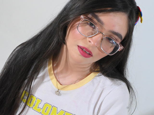 EvaMcAllister with long black hair, pink lipstick, and clear glasses, wearing a yellow-trimmed white shirt, has a slight smirk, and a small pendant on a necklace.