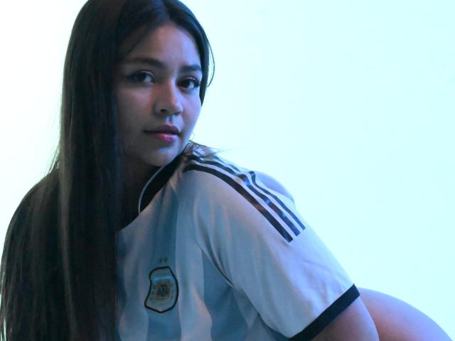 KimDevinne, con cabello castaño largo, vistiendo una camiseta blanca y azul de fútbol argentino, mirando por encima del hombro, con una luz azulada, mostrando su trasero..