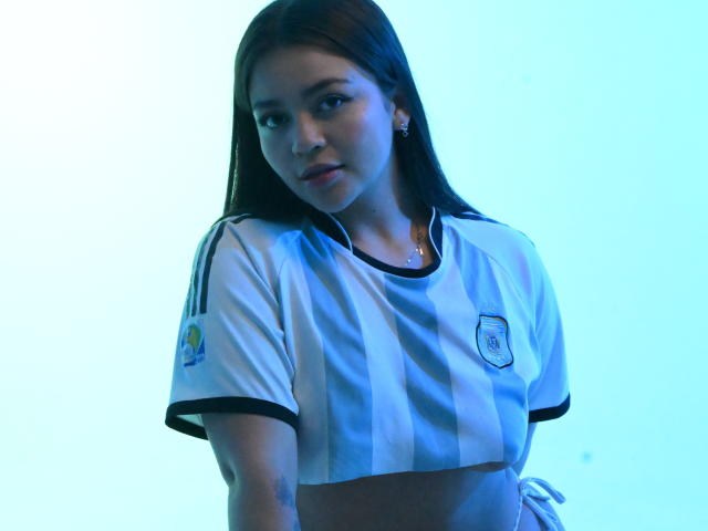 KimDevinne, con cabello castaño largo, vestida con una camiseta de fútbol a rayas blancas y azules, que deja ver sus pechos, iluminación azul y una ligera sonrisa, mirando a la cámara..