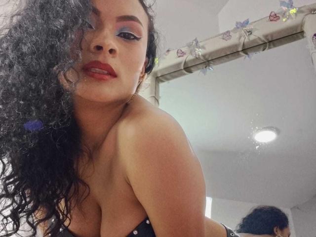 Kalie Bennett, met krullend zwart haar, rode lippenstift en blauwe oogmake-up, leunt voorover, toont haar borsten en kijkt in een spiegel met vlinderdecoraties op de lijst..