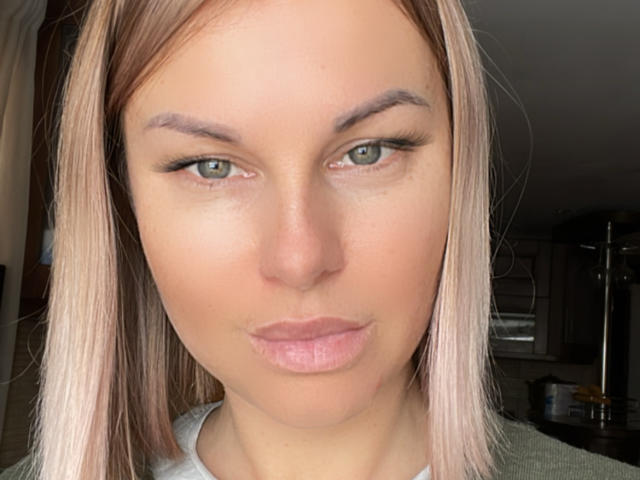 BelleLisaG tiene ojos verde claro, labios rosados y cabello rubio lacio, con un ligero bostezo, y lleva una camisa verde. Su rostro está centrado y mira directamente a la cámara..