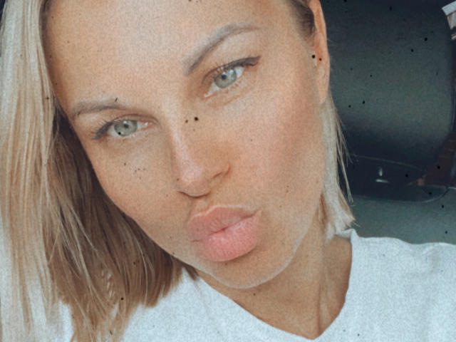 BelleLisaG's pouty lips, light blonde bob, and green eyes are front and center. She's got a few dark moles on her face and is wearing a white tee.