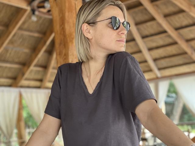 BelleLisaG avec des cheveux blonds courts, portant un t-shirt noir à col en V et des lunettes de soleil réfléchissantes, regardant sur le côté avec une expression calme, sous un toit en bois..