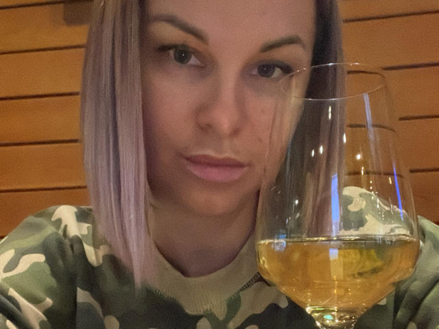 BelleLisaG con capelli biondo-rosati, che indossa un maglione verde mimetico, tiene in mano un bicchiere di vino bianco, con le labbra leggermente serrate e guarda direttamente la fotocamera..