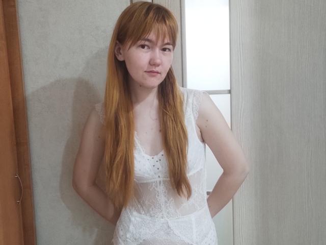 SheritaChieng with long red hair, wearing a white lace dress, standing with hands on hips, showing small tits and a slim, pale, pretty face with a slight smile.