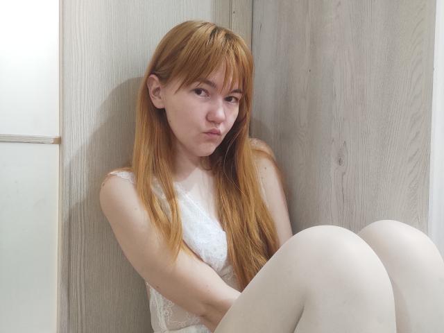 SheritaChieng hat langes, rotes Haar, blasse Haut und kleine Brüste, sitzt mit angezogenen Knien, trägt ein weißes Spitzenoberteil und weiße Strumpfhosen und sieht leicht schmollend aus..
