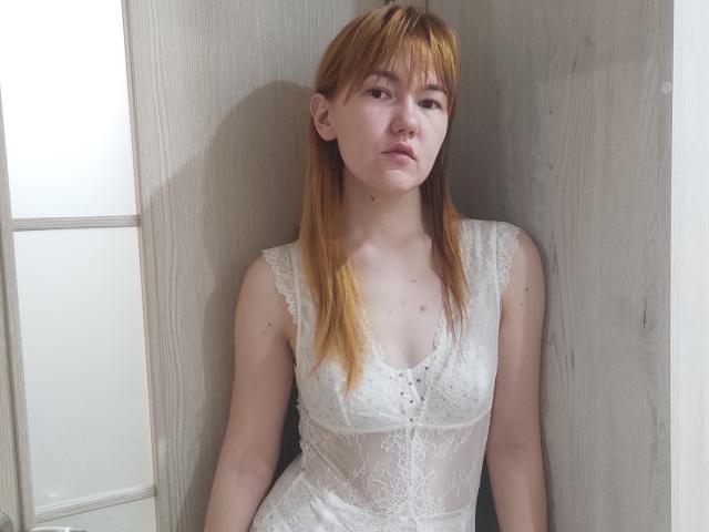 SheritaChieng, con piel pálida, cabello naranja y senos pequeños, vestida con un vestido blanco de encaje, sentada contra una pared de madera, mirando ligeramente hacia la izquierda con una expresión neutra..