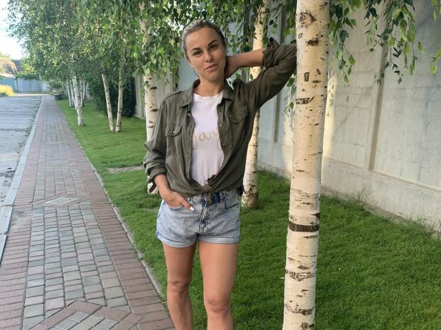 BelleLisaG con piel morena, camisa verde atada, camiseta blanca y pantalones cortos vaqueros azules, apoyada en un abedul, con una mano en el bolsillo, hierba verde y un camino de ladrillos en el fondo..
