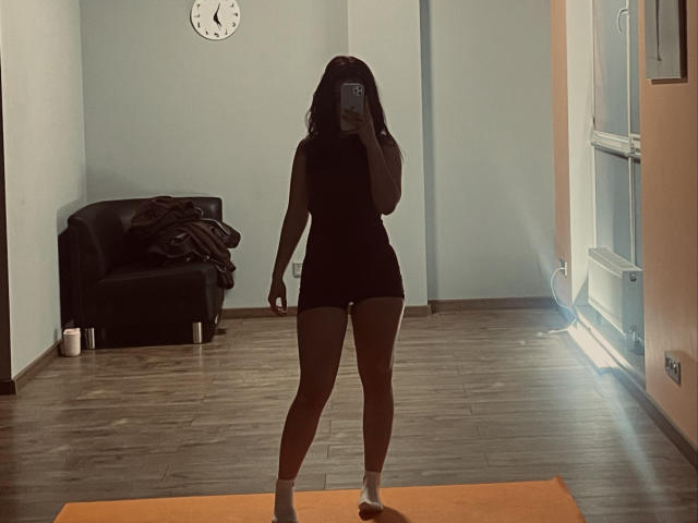 AlexXas is standing on an orange mat, wearing a black sleeveless top and shorts, holding a phone, covering her face, with her ass and pussy slightly visible.