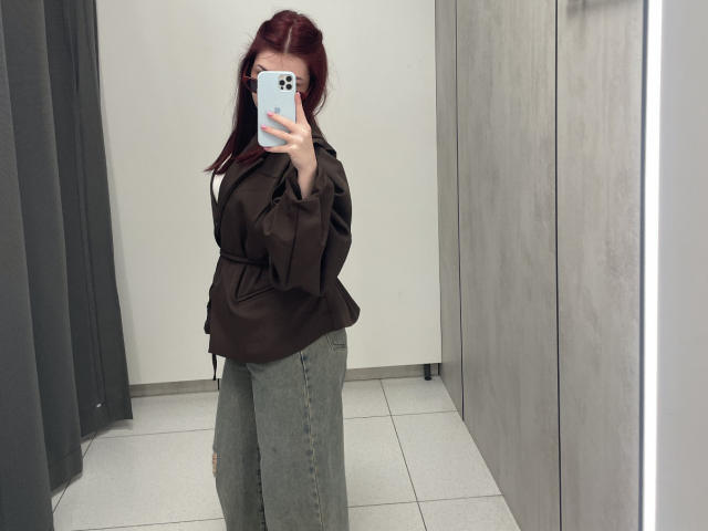 AlexXas with red hair, brown jacket, and green pants, taking a mirror selfie, showing her phone-covered face, in a gray-walled, tiled room.