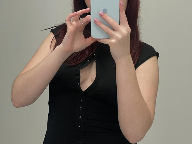 AlexXas with medium tits, black button-up top, holding a white iPhone, covering face, pink nails, long dark brown hair, fair skin, grey background, taking a selfie.